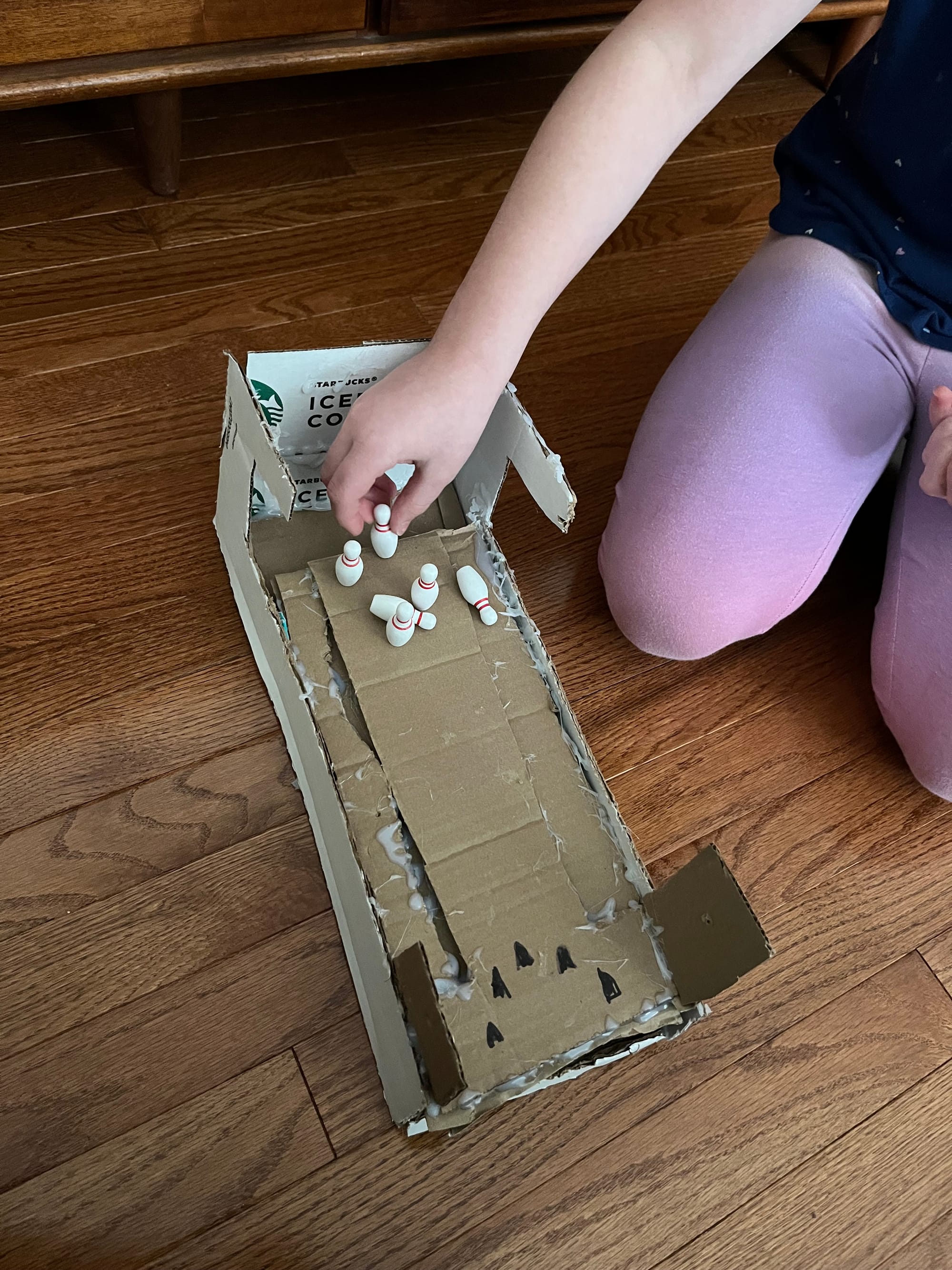 Rustic homemade mini bowling alley, made of cardboard scraps and hot glue with tiny bowling pins. Child's knees and hands visible in background.
