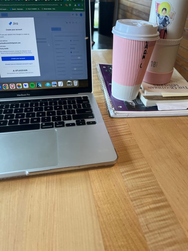 Laptop open to organization software. Stack of notebook, books, disposable pink and white coffee cup, and pink and white water bottle with watercolor Joan of Arc sticker.
